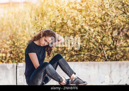 Sportliche Frau, die auf dem Boden sitzt. Die Hand hinter dem Kopf. Hintergrund bei Sonnenuntergang Stockfoto