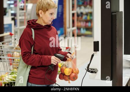 Blonde, reife Konsumentenin der Freizeitkleidung, die sich den Produktpreis auf dem Bildschirm ansieht, während sie die Packung frischer reifer Äpfel im Supermarkt scannt Stockfoto