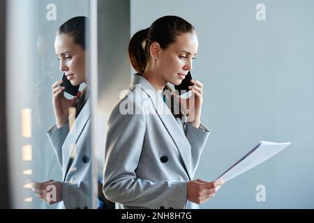 Junge Finanzdirektorin in grauem Anzug, die Vertragspunkte durchsucht und mit dem Geschäftspartner über ein Mobiltelefon im Büro spricht Stockfoto