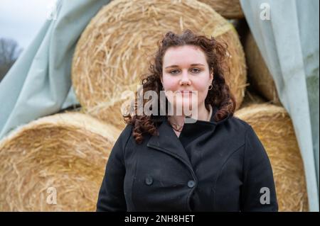 Eine Frau mit braunen Locken steht vor gestapelten Strohballen, lächelt und schaut in die Kamera, bewölkter Tag Stockfoto