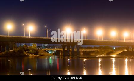 Nachtstraße mit Laternen, die von der Bogenbrücke in einen Fluss mit Reflexionen in Russland führt. Stockfoto