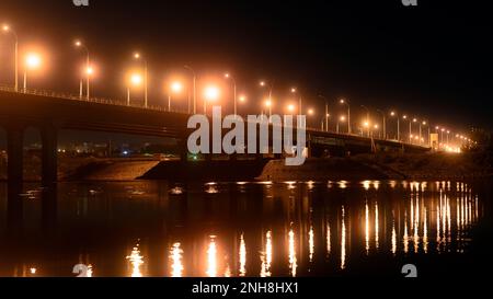 Nachtstraße mit Laternen, die von der Bogenbrücke in einen Fluss mit Reflexionen in Russland führt. Stockfoto