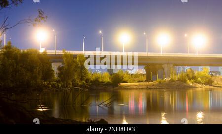 Nachtstraße mit Laternen, die von der Bogenbrücke in einen Fluss mit Reflexionen in Russland führt. Stockfoto