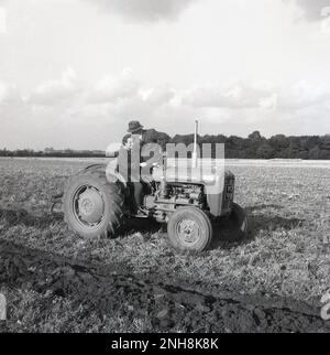 1965, historisch, Bauer, in Hemd und Krawatte, Jacke und Hut auf einem Traktor neben einer Fahrerin, Barton, England, Großbritannien. Die Frau hat eine Lektion, wie man ein Feld pflügt. Der Traktor ist ein Massey-Ferguson 35X, ein klassisches Modell der Epoche. Stockfoto