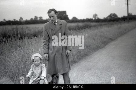 1950er, historisch, ein junger Vater, ein Hemd und eine Krawatte und ein Trenchcoat, laufen mit seiner kleinen Tochter und seinem Hund draußen auf einer Landstraße, das kleine Mädchen reitet auf einem kleinen Dreirad, England, Großbritannien. Stockfoto