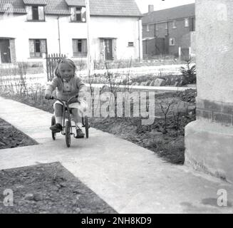 1950er, historisch, eine kleine Kippe, die auf ihrem Dreirad auf einem kürzlich erbauten Betonweg auf einem neuen Wohngebiet in England, Großbritannien, fährt. Stockfoto