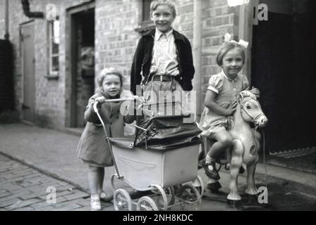 1950er, historisch, drei glückliche kleine Kinder, Bruder mit seinen Schwestern, draußen in einer Kopfsteinpflasterstraße, am Eingang zu ihrem Zuhause, England, Großbritannien. Die kleinen Mädchen sind mit ihren Spielzeugen da, eine mit ihrem Puppenwagen - einer aus Metall gefertigten Version für Minibusse - und das andere Mädchen sitzt auf ihrem Spielzeugpferd. Stockfoto