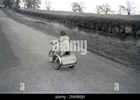 1950er, historisch, ein kleines Mädchen mit Mantel und Wollmütze, das auf einem stahlgerahmten Dreirad mit Stiefel entlang einer von Hecken gesäumten Landstraße in England, Großbritannien, fährt. Stockfoto