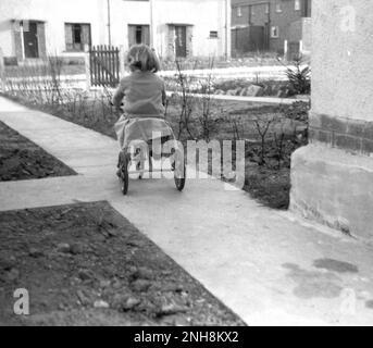 1950er, historisch, Blick von hinten auf eine kleine Kiemen, die auf einem kürzlich erbauten Betonweg auf einem neuen Wohngebiet in England, Großbritannien, auf ihrem Dreirad reitet. Stockfoto