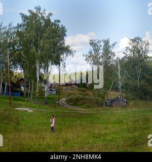 Ein russisches Mädchen geht über ein Feld in Richtung einer Straße und Häuser auf einem Hügel in der Nähe eines Waldes im Dorf Sibirien. Stockfoto