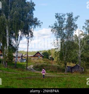 Ein russisches Mädchen geht über ein Feld in Richtung einer Straße und Häuser auf einem Hügel in der Nähe eines Waldes in Sibirien. Stockfoto