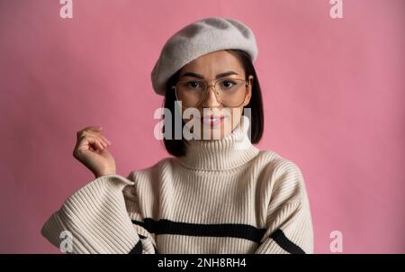 Wunderschöne braune Frau in gestreiftem Pullover und Baskenmütze, stehend und in der Kamera, isoliert auf Pink Stockfoto