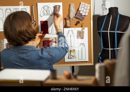 Eine Designerin arbeitet mit Skizzen und Stoffproben, um Kleidung an ein Korkbrett zu nähen. Stockfoto