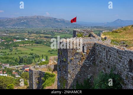 Albanische Flagge auf den Burgmauern von Schloss Rozafa/Kalaja e Rozafës und Blick auf die albanischen Alpen in der Nähe von Shkodër/Shkodra im Nordwesten Albaniens Stockfoto