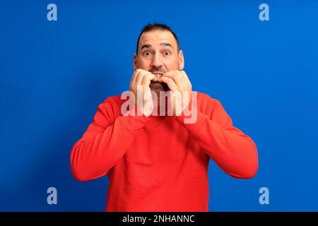 Ein hübscher Mann mit rotem Pullover auf isoliertem blauem Hintergrund, der gestresst und nervös aussieht, mit Händen am Mund, die Nägel kauen. Angstproblem Stockfoto
