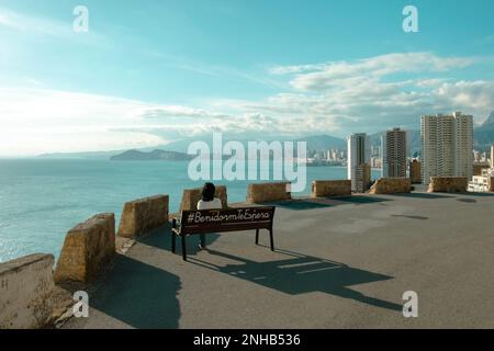 Benidorm Wanderroute Punta del Cavall, Naturpark, Torre Punta del Cavall. Frau sitzt auf einer Bank mit Inschrift Benidorm te espera und bewundert Stockfoto