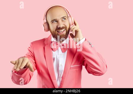 Junger Mann in pinkfarbenem Partyanzug, Fliege und Kopfhörern, der Musik hört und lächelt Stockfoto