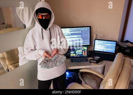Ein Hacker mit Geld. Internetdiebstahl. Ein Mann, der eine Balaklava hinter einem Laptop trägt. Stockfoto