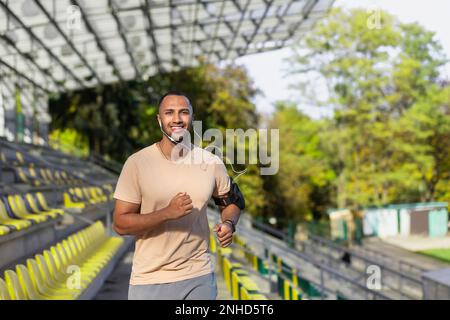 Junger afroamerikanischer Mann, Sportler, Leichtathlet, der im Sommer im Stadion zwischen den Tribünen mit Kopfhörern läuft. Sie lächelte in die Kamera. Stockfoto