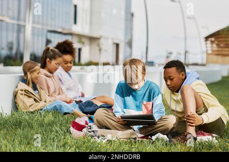 Zwei interkulturelle Schuljungen, die auf einem Tablet-Bildschirm sitzen und auf einem grünen Rasen im Park sitzen, gegen eine Gruppe erholsamer Mädchen, die Gadgets verwenden Stockfoto