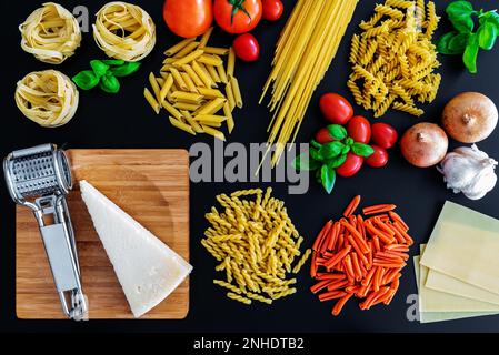 Flacher Lay von verschiedenen ungekochten italienischen Nudeln auf dunklem Hintergrund mit Basilikumblättern, frischen Tomaten und Zwiebeln, Parmesan-Käse und Reibe Stockfoto