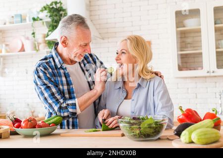 Älterer Mann, der Gurke füttert, schneidet ihre Ehefrau in die Küche Stockfoto