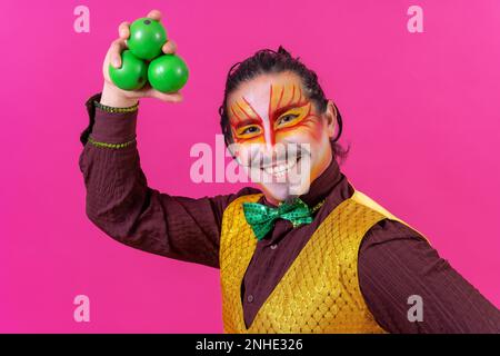 Clown mit weißer Gesichtsschminke auf pinkfarbenem Hintergrund, jonglierende grüne Bälle Stockfoto