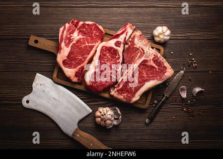 Set aus drei verschiedenen Arten roher Fleischsteaks: Ribeye, T-Bone, Cowboy auf Schneidebrett mit Messer und Gewürzen, Holzhintergrund. Gereifte Steaks Stockfoto