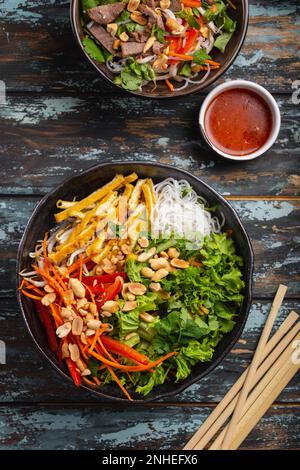 Nudelsalat im asiatischen, chinesischen oder vietnamesischen Stil mit frischem Gemüse, gebratenem Tofu und Erdnüssen, serviert in einer rustikalen Keramikschüssel auf farbenfrohem Holz Stockfoto