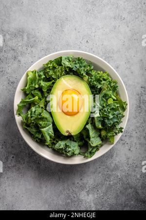 Salatschüssel mit Grünkohl für ausgewogenes Menü, rohes Eigelb in frisch geschnittener halber Avocado, reich an gesunden Fetten auf grauem Steinhintergrund. Ketogene kohlenhydratarme Stockfoto