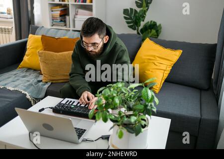 Mann, der elektronische Musiktitel mit tragbarer MIDI-Tastatur auf einem Laptop im Heimstudio aufnimmt. Produzieren und Mischen von Musik, die Rhythmen erzeugt und Stockfoto