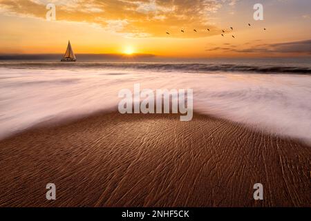 Ein Segelboot segelt entlang des Ozeans mit Wellenbrechern an der Küste Stockfoto