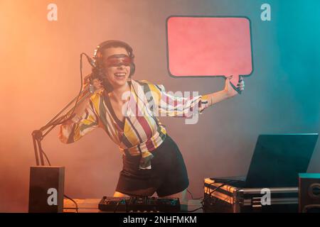 Junger Musiker hält eine Sprechblase mit Papppapier, um eine Nachricht auf einer isolierten Mustervorlage zu veröffentlichen. DJ-Frau, die Ikone auf Plakatwänden zeigt und Techno-Musik im Studio mit Rauch mischt. Stockfoto