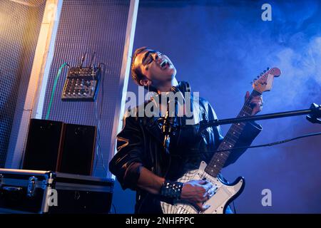 Cooles Sängermädchen, das Heavy-Metal-Musik mit Gitarre spielt, am Mikrofon singt und sich ekstatisch fühlt. Bass-Instrument spielen, um alternative Rock-Live-Auftritte im Studio zu machen, in Lederbekleidung. Stockfoto