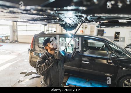 Mittelgroße Aufnahme eines Mechanikers, der das Fahrgestell inspiziert. Noch ein Auto im Hintergrund. Hochwertiges Foto Stockfoto