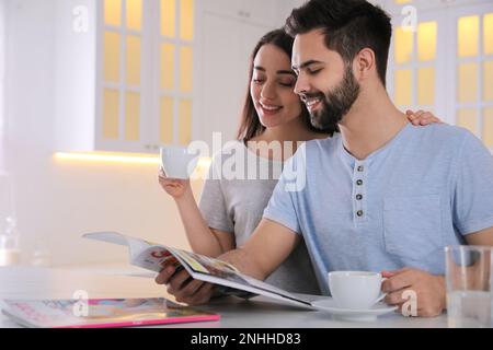 Ein junges Paar mit Tassen Getränke, das eine Zeitschrift am Tisch in der Küche liest Stockfoto