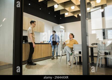 Gruppe fröhlicher junger Menschen, die während der Kaffeepause im Büro sprechen und lächeln. Team professioneller Softwareentwickler, das sich im entspannt Stockfoto