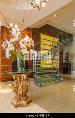 Glasvase mit weißen Seidenblumen auf dem Sockel der chinesischen Löwenfigur mit Glasplatte sowie gedrehten Treppen aus Stahl und gehärtetem Glas. Stockfoto