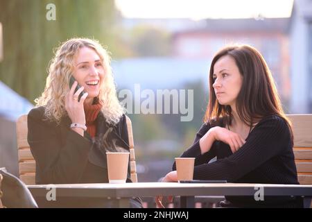 Glückliche junge Frau, die auf dem Handy chattet und ihre gelangweilte Freundin ignoriert. Weibliche Freunde sitzen im Straßencafe im Freien und haben harte Zeit zu kommunizieren Stockfoto