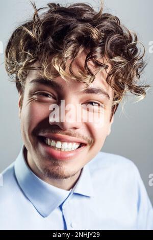 Portrait eines fröhlichen, lächelnden, gutaussehenden Mannes mit Schnurrbart, lockiger Frisur, blauem Hemd, der in die Kamera schaut, Glück ausdrückt. Studioaufnahmen im Innenbereich isoliert auf grauem Hintergrund. Stockfoto