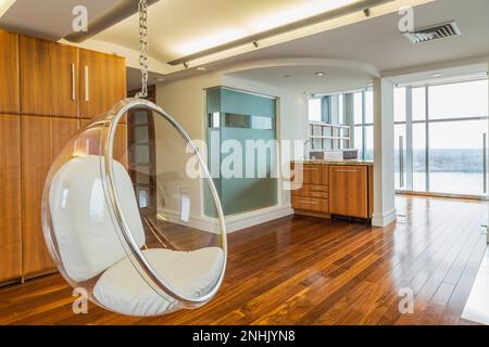 Klarer, halbrunde Plastikstuhl hängt von der Decke im Heimbüro mit exotischen Holzfußböden auf der obersten Etage in einer modernen, luxuriösen Wohnung. Stockfoto