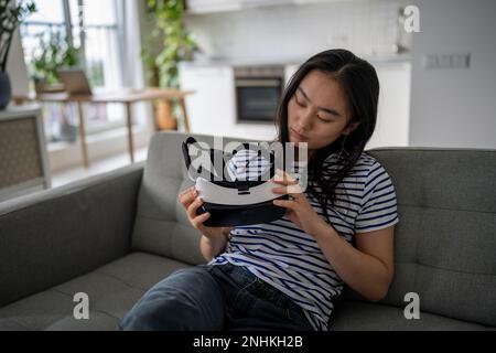 Ein fokussiertes asiatisches Mädchen sitzt auf dem Sofa zu Hause und schaut sich eine Virtual-Reality-Brille für ein Smartphone an, das von einem Freund gegeben wurde Stockfoto