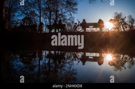 München, Deutschland. 21. Februar 2023. Die Sonne spiegelt sich in den Abendstunden in einem kleinen Bach wider, an dessen Ufern einige Wanderer die Frühlingstemperaturen genießen. Die Monopteros sind auf der linken Seite zu sehen. Kredit: Peter Kneffel/dpa/Alamy Live News Stockfoto