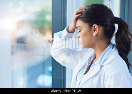 Es ist ein stressiger Beruf. Eine junge Ärztin sieht gestresst aus, während sie in einem Krankenhaus am Fenster steht. Stockfoto