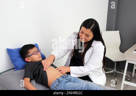 Eine lateinische Ärztin prüft den Bauch eines Kindes in ihrer Praxis, um Krankheiten wie Blinddarmentzündung, Gastroenteritis, Hernie oder Geschwür auszuschließen Stockfoto