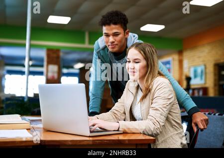Wir helfen uns gegenseitig bei der Vorbereitung auf die Prüfungen. Ein junger Mann schaut zu, während eine Studentin an einem Laptop arbeitet. Stockfoto