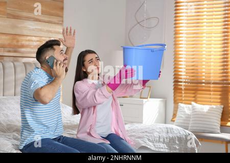 Eine junge Frau sammelt Wasser von der Decke, während ihr Freund zu Hause den Dachreparaturservice anruft Stockfoto