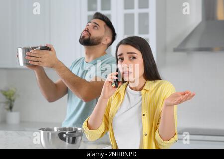 Ein junger Mann sammelt Wasser aus der Decke, während seine Freundin den Dachreparaturservice in der Küche anruft Stockfoto