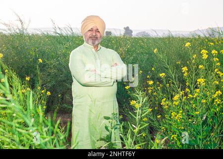 Porträt eines lächelnden Senior Punjabi sikh Mannes, der pagdi und traditionelle Kurta-Outfits trägt und auf dem Landwirtschaftsfeld quer über die Arme steht. Stockfoto