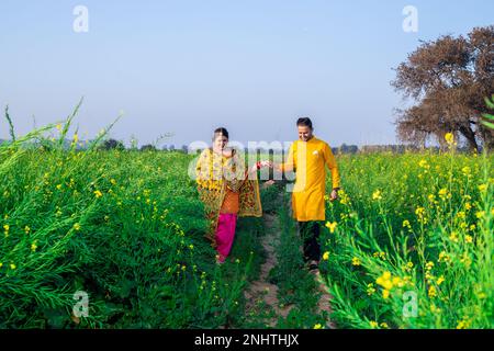 Porträt eines glücklichen jungen indischen punjabi-Paares, das farbenfrohe Outfits trägt und sich gegenseitig die Hand hält, wenn er auf dem Landwirtschaftsfeld zusammen geht. Stockfoto